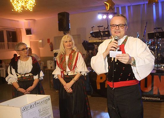 Ein Mann hält ein Mikrofon und liest ein Stück Papier, neben zwei Frauen in traditioneller Kleidung. Dahinter befindet sich eine Bühne mit Musikinstrumenten und einem Hintergrund, auf dem 'PUN' steht. Ein Kasten mit 'QUIZVERLOSUNGS' ist im Vordergrund.