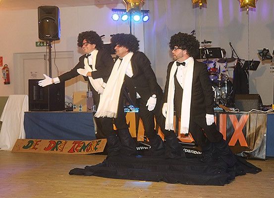 Drei Männer in Anzügen mit weißen Schals führen eine Darbietung auf der Bühne auf. Hinter ihnen steht ein Tisch mit einer weißen Tischdecke und einem Schild. Im Hintergrund sind Musikinstrumente und Lautsprecher aufgestellt.