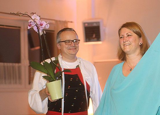Ein Mann mit Brille und einer Schürze hält eine Orchidee in einem Topf, während er neben einer lächelnden Frau in einem türkisen Oberteil steht.
