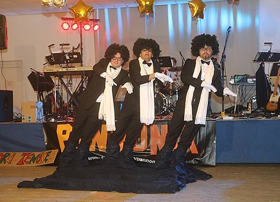 Drei Personen in Anzügen mit weißen Schals und Fliegen stehen auf der Bühne mit einem Mikrofon, Lautsprechern und Musikinstrumenten im Hintergrund.