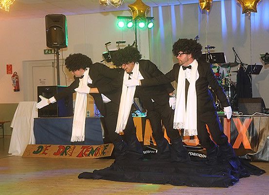 Drei Männer führen auf einer Bühne auf, tragen weiße Schals und schwarze Anzüge mit Fliegen. Sie stehen auf einem schwarzen Tuch. Dahinter sind ein Banner mit roten Buchstaben und Musikinstrumente zu sehen.