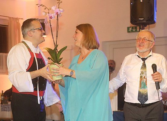Drei Personen stehen auf der Bühne; eine Frau hält eine Pflanze, ein Mann ein Mikrofon und ein Mann mit Brille einen Topf mit Blumen.