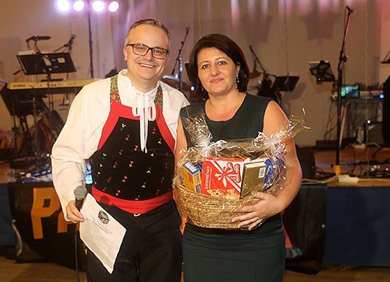 Ein Mann und eine Frau stehen lächelnd zusammen auf der Bühne. Der Mann hält ein Mikrofon und ein Papier, die Frau einen Geschenkkorb. Dahinter steht eine Bühne mit Musikinstrumenten und einem Hintergrund.