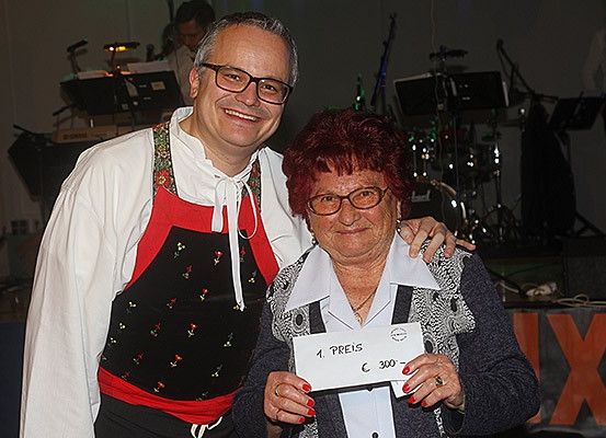 Ein Mann und eine Frau stehen lächelnd da, wobei die Frau ein Schild mit der Aufschrift 1 Preis und 300 € hält. Sie stehen vor einer Bühne mit Musikinstrumenten und einem Banner mit dem Buchstaben X.