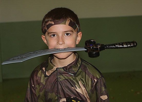 Ein junger Junge in einer Tarnjacke und einem Stirnband hält ein Schwert im Mund.