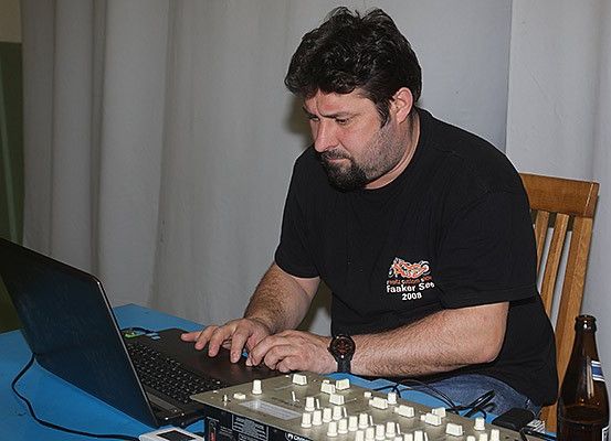 Ein Mann sitzt an einem Tisch mit Laptop und Tonequipment, konzentriert auf den Bildschirm. Er trägt ein schwarzes T-Shirt und hat eine Uhr am Handgelenk. Hinter ihm befindet sich ein Vorhang und eine Bierflasche.