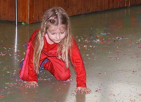 Ein kleines Mädchen mit geflochtenen Haaren kniet auf einem mit buntem Konfetti bedeckten Boden. Sie trägt ein rotes Outfit.