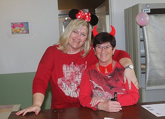 Zwei Frauen in roten Shirts posieren für ein Foto. Eine trägt eine Mickey-Mouse-Kopfband und die andere ein Teufelshorn-Kopfband. Beide lächeln. Eine Flasche steht auf dem Tisch. Ein Kühlschrank und ein Ballon sind im Hintergrund.