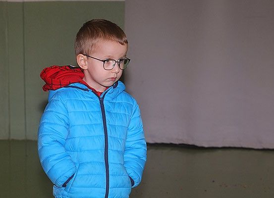 Ein junger Junge in einem blauen Mantel mit roter Kapuze steht drinnen und schaut mit einer Brille nach unten. Der Raum hat eine grüne Wand und einen weißen Hintergrund.