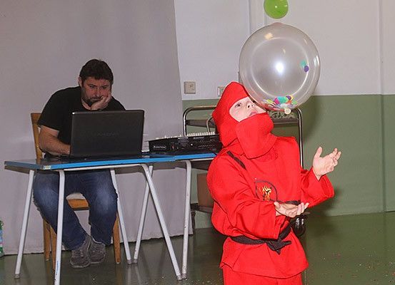 Ein Mann sitzt an einem Schreibtisch mit Laptop, während ein Kind in einem roten Kostüm einen großen, bunten Ballon hält.