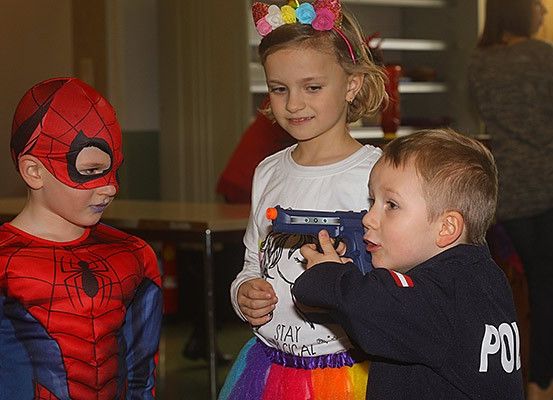Ein Junge in einem Spiderman-Kostüm und ein Mädchen mit weißer Bluse und Blumen-Haarband spielen mit einer Spielzeugpistole. Eine Frau steht im Hintergrund.