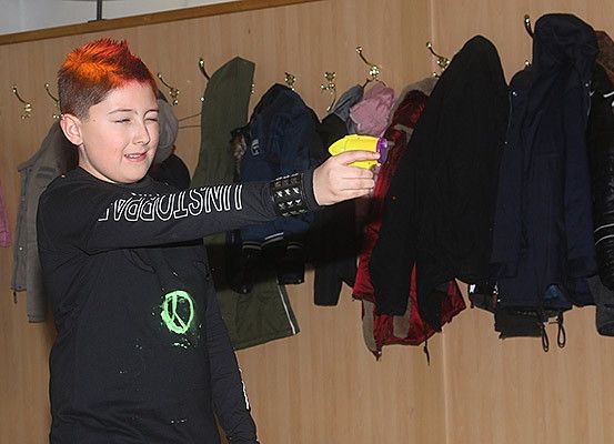 Ein junger Junge mit roten Haaren, der ein schwarzes T-Shirt trägt, hält eine gelbe und lila Spielzeugpistole vor einer Wand mit vielen hängenden Jacken.