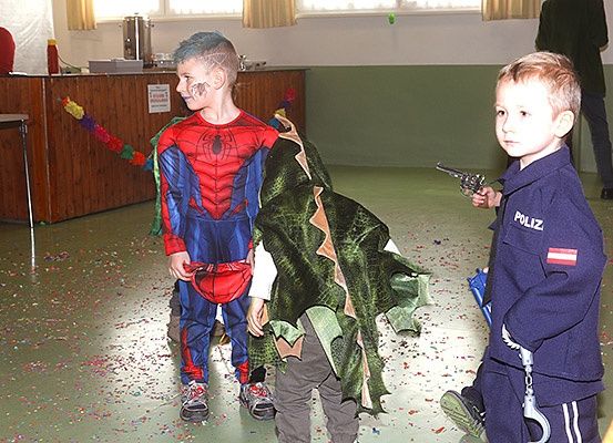 Zwei Kinder stehen in einem Raum mit Konfetti auf dem Boden. Ein Kind ist als Spiderman verkleidet, das andere als Polizist. Dahinter steht eine Holztheke mit einer Kaffeemaschine und einer Blumendekoration.