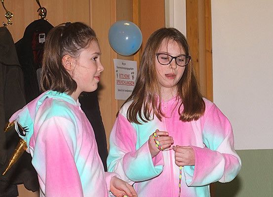 Zwei Mädchen in rosa und weißen Tie-Dye-Kleidern stehen in einem Raum. Ein Mädchen trägt eine Brille und hält ein Armband. Sie schauen sich etwas an. Hinter ihnen ist ein blauer Ballon an einer Tür mit einem Schild befestigt.