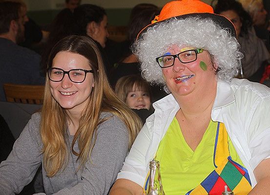 Zwei Frauen, eine in einem bunten Kostüm, sitzen an einem Tisch in einem Restaurant, während andere Personen um sie herum sitzen und stehen.
