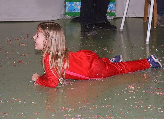 Ein kleines Mädchen in einem roten Outfit liegt auf einem mit buntem Konfetti bedeckten Boden. Hinter ihr stehen zwei Personen in dunklen Hosen und Schuhen in der Nähe eines Tisches.