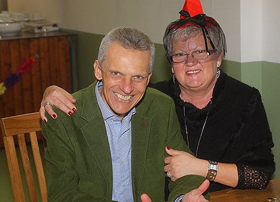 Ein älterer Mann und eine ältere Frau sitzen in einem Raum und lächeln. Der Mann trägt eine grüne Jacke und die Frau trägt eine Brille und einen roten Hut. Hinter ihnen steht eine Holztheke mit einer Schüssel und einem Teller.