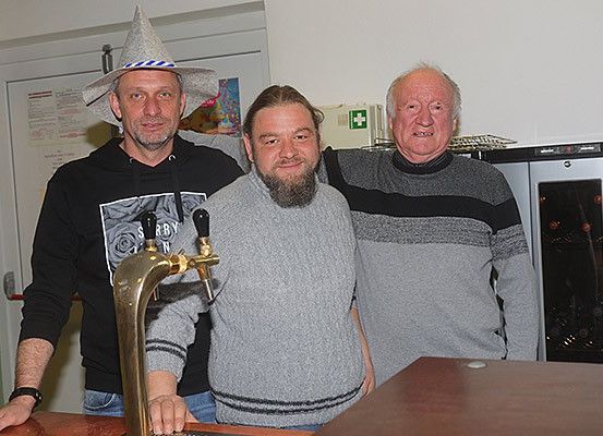 Drei Männer stehen zusammen in einem Raum. Der Mann auf der linken Seite trägt eine schwarze Kapuzenjacke, der in der Mitte ein graues Sweatshirt und der auf der rechten Seite ein gestreiftes Sweatshirt. Hinter ihnen befindet sich eine weiße Wand mit einer Tür.