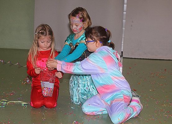Drei junge Mädchen spielen auf dem Boden, eine hält eine Plastiktüte mit Konfetti. Die Mädchen haben Gesichtsbemalung.
