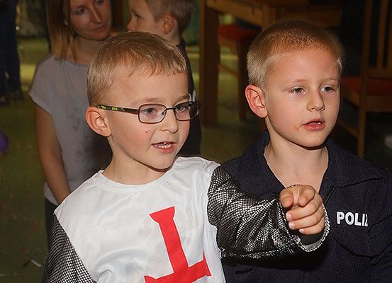 Zwei Jungen in Kostümen stehen nebeneinander, einer mit einem roten Kreuz auf seinem Shirt und der andere zeigt auf etwas. Hinter ihnen sind eine Frau und ein Kind anwesend.