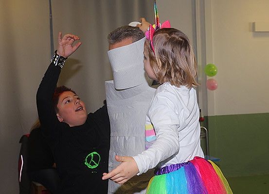 Ein junger Junge in einem schwarzen T-Shirt tanzt mit einem Mädchen in einem Einhornkostüm, während ein Mann in einer Papiermaske zusieht. Die Umgebung ist drinnen mit Luftballons und einer grünen Wand.