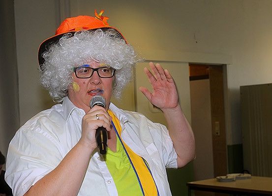 Eine Person mit Perücke und Brille hält ein Mikrofon und scheint eine Rede zu halten.