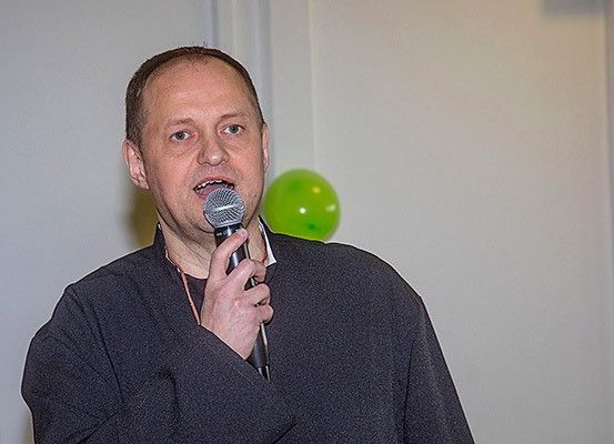 Ein Mann steht mit einem Mikrofon und spricht zu einer weißen Wand, mit einem grünen Luftballon im Hintergrund.