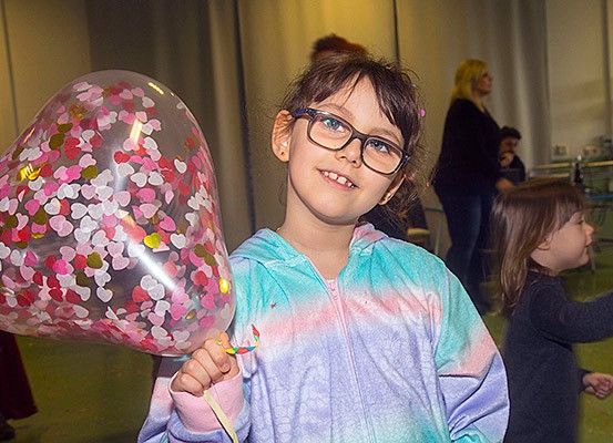 Ein junges Mädchen mit Brille hält einen herzförmigen Luftballon mit Konfetti. Sie lächelt und steht in einem Raum mit anderen Kindern.