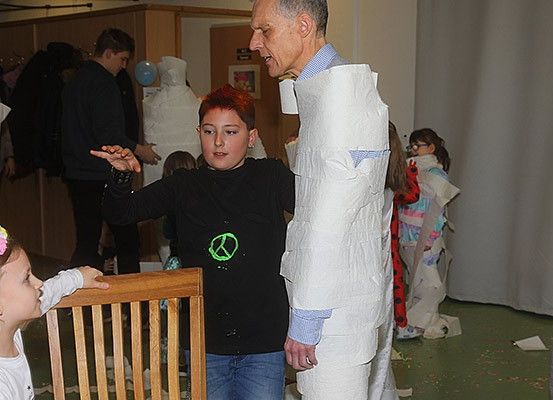 Ein in Toilettenpapier gewickelter Mann spricht mit einem Jungen, dessen T-Shirt ein Friedenszeichen trägt, während ein Mädchen zusieht, in einem Raum mit anderen Kindern und einem weißen Vorhang.