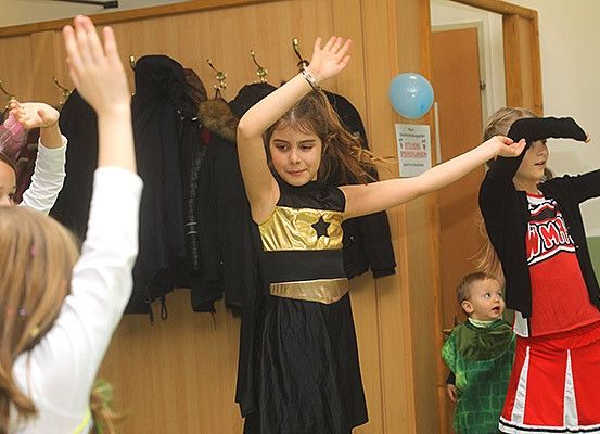Kinder in Kostümen, darunter eines mit einem Stern auf ihrem Kleid, tanzen in einem Raum mit Holzwänden und hängenden Jacken im Hintergrund. Ein blauer Ballon ist im Hintergrund zu sehen.