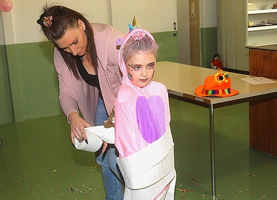Eine Frau hilft einem jungen Mädchen in einem Einhornkostüm mit einer Rolle Toilettenpapier auf einem grünen Boden. Dahinter steht ein Tisch mit einem Hut mit einer Sonnenblume.