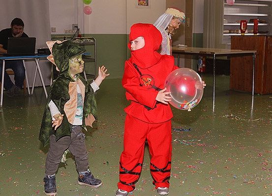 Zwei Kinder in Kostümen spielen in einem Raum, eines hält einen bunten Ballon. Ein Mann sitzt auf einem Stuhl mit einem Laptop, und eine andere Person steht hinter einem Tisch.