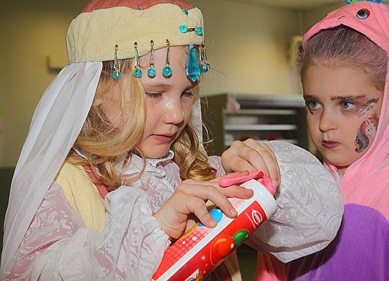 Zwei Mädchen spielen mit einer Tube Zahnpasta. Das Mädchen auf der linken Seite trägt eine Kopfbedeckung mit blauen Kristallen. Das Mädchen auf der rechten Seite trägt ein rosa Einhornkostüm.