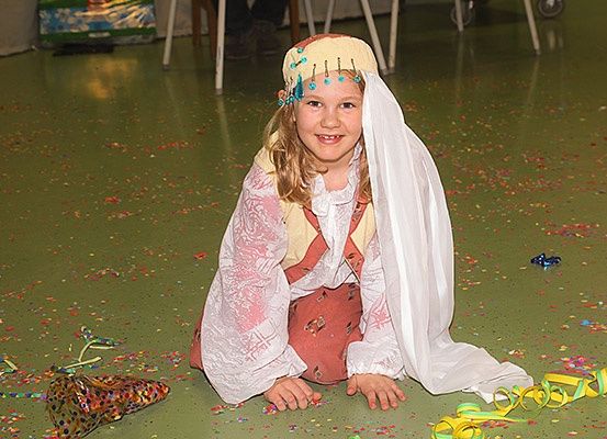 Ein kleines Mädchen in traditioneller Kleidung sitzt auf dem Boden, umgeben von Konfetti, wahrscheinlich während einer festlichen Veranstaltung.