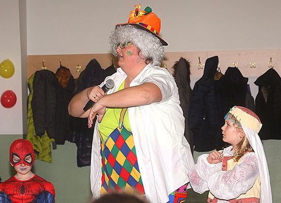 Ein Clown in einem Kostüm hält ein Mikrofon in der Hand, neben ihm steht ein Kind in einem Spider-Man-Kostüm und ein weiteres in einem traditionellen Kleid. Dahinter hängen Kleider an Haken und Ballons an der Wand.