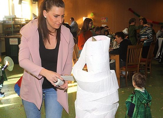 Eine Frau steht neben einem großen Stapel Toilettenpapier und hält eine Rolle in der Hand, während Kinder in Kostümen um sie herum spielen. Hinter ihr sitzen Menschen an Tischen, einige von ihnen tragen Brillen.