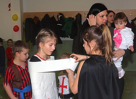 Mehrere Kinder in Kostümen, darunter Ritter, versammeln sich um eine Frau, die ein Baby hält. Ein Kind wickelt eine Rolle Toilettenpapier um einen anderen, während andere zuschauen und einige Jacken im Hintergrund hängen.