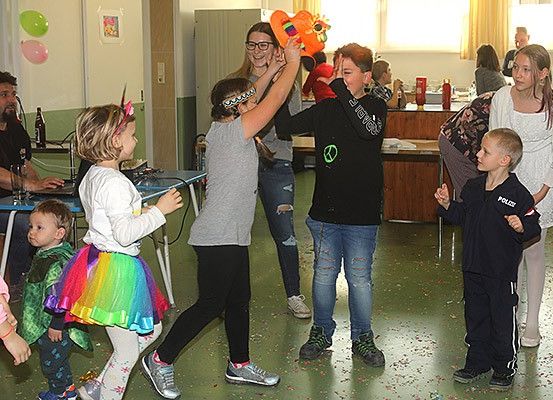 Mehrere Kinder und Erwachsene befinden sich in einem Raum. Ein junges Mädchen hält einen orangefarbenen Ballon und bläst ihn auf. Die Kinder spielen im Raum herum.