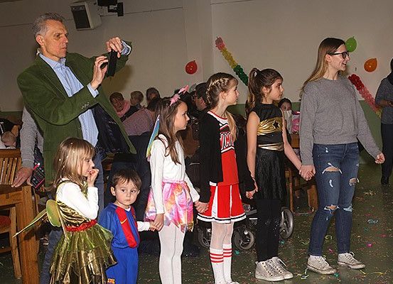 Eine Gruppe von Kindern in Kostümen, darunter eine Fee und Superhelden, stehen in einem festlichen Raum mit Luftballons und Girlanden zusammen mit Erwachsenen. Ein Mann macht ein Foto.