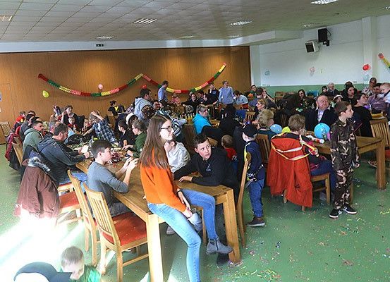 Ein Raum voller Menschen, wahrscheinlich für eine Party. Einige sitzen an Tischen, während andere stehen und sich unterhalten. Bunte Dekorationen und Ballons sind vorhanden.