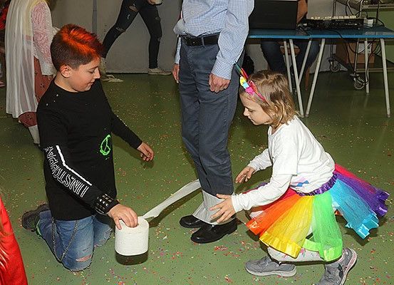 Ein junger Junge mit roten Haaren zieht eine Rolle Toilettenpapier, während ein Mädchen in einem Tutu es hält. Ein Mann steht hinter ihnen und andere sind im Hintergrund.