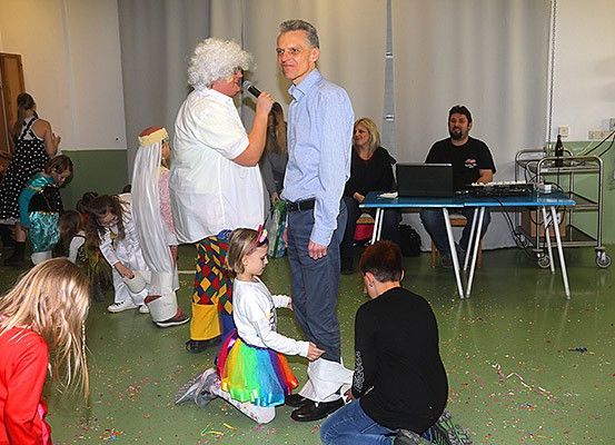 Eine Gruppe von Menschen ist in einem Raum, einige tragen Kostüme, und ein Mann spricht in ein Mikrofon. Einige sitzen auf dem Boden, während andere stehen. Ein Tisch mit einem Laptop ist in der Nähe.
