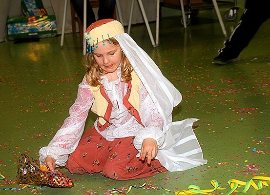 Ein junges Mädchen in traditioneller Kleidung sitzt auf einem mit Konfetti bedeckten grünen Boden. Sie hält einen Schuh.