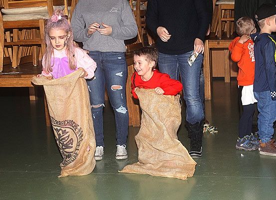 Zwei Kinder spielen Sackhüpfen in einem Saal und tragen Säcke mit etwas darin. Hinter ihnen beobachten andere Kinder.
