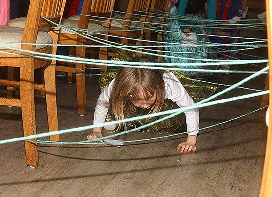 Ein kleines Mädchen navigiert durch ein Labyrinth aus blauen Schnüren auf einem Holzboden, während andere auf Stühlen sitzen.