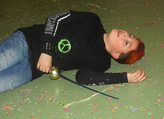 Ein junger Mensch liegt auf einem mit Konfetti bedeckten grünen Boden, trägt ein schwarzes Shirt mit einem Friedenszeichen. Sie halten eine goldene Kugel und ein Schwert.