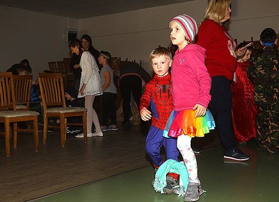 Zwei Kinder in Kostümen gehen durch einen Flur, umgeben von Erwachsenen und Kindern. Der Raum hat Holzstühle und einen grünen Boden.