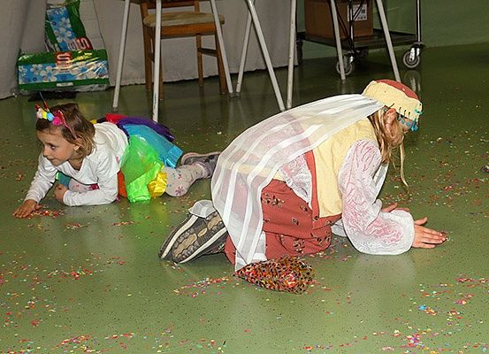 Zwei Kinder spielen auf einem grünen Boden mit Konfetti, eines ist in einem bunten Rock gekleidet und das andere in einem Kostüm mit Kopfbedeckung. In der Nähe stehen Stühle, ein Tisch und eine Kiste.