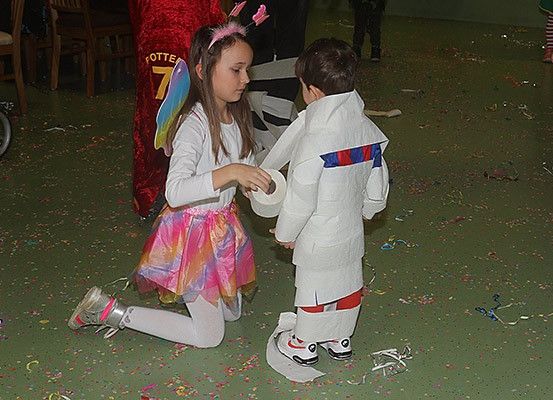 Zwei Kinder spielen in einem Raum mit Konfetti. Ein Mädchen in einem rosa Kostüm kniet und hält eine Rolle Papier, während ein Junge in einem weißen Kostüm steht. Hinter ihnen ist eine Person in einem roten Kostüm zu sehen.