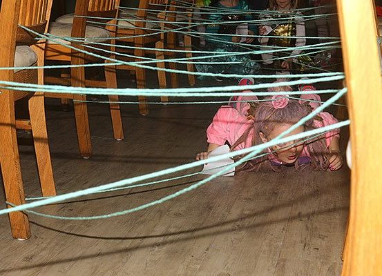 Ein kleines Mädchen navigiert durch ein Labyrinth aus grünen Seilen auf einem Holzboden, hält ein Stück Papier in der Hand, mit Stühlen und einem anderen Kind im Hintergrund.